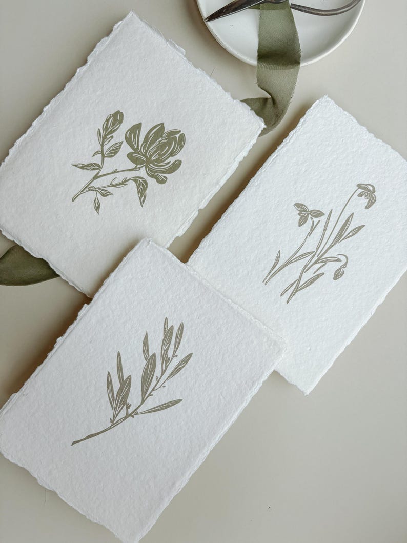 May include: Three white handmade paper cards with botanical illustrations in olive green. Each card features a different floral or plant design, including a flower, sprigs, and leaves. The cards have deckled edges.