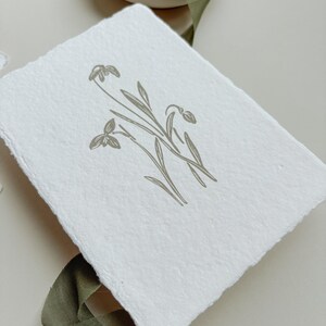 May include: A white, textured paper with a botanical illustration of snowdrop flowers in a muted green hue. The artwork features delicate lines and a minimalist design, evoking a sense of natural elegance. A green ribbon and scissors are in the background.