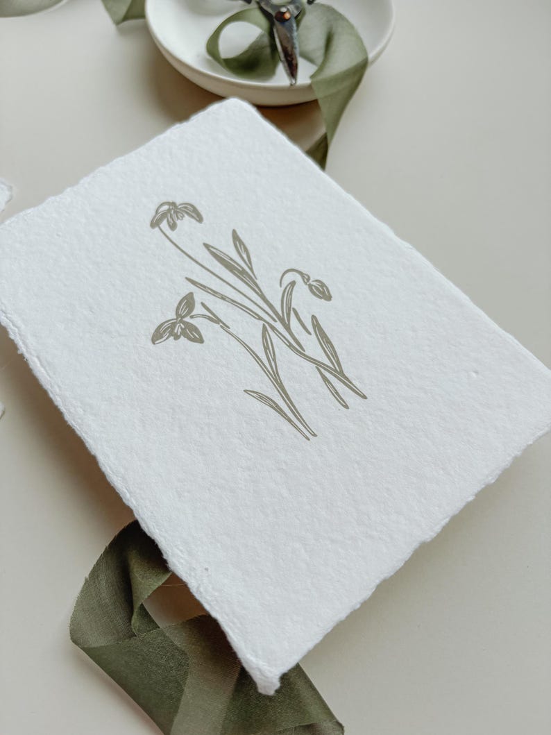 May include: A white, textured paper with a botanical illustration of snowdrop flowers in a muted green hue. The artwork features delicate lines and a minimalist design, evoking a sense of natural elegance. A green ribbon and scissors are in the background.