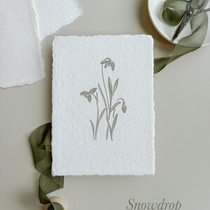 May include: A white, textured card with a botanical illustration of snowdrop flowers in a muted green hue. The word "Snowdrop" is printed in a matching color. A green ribbon, scissors, and additional paper are arranged around the card.