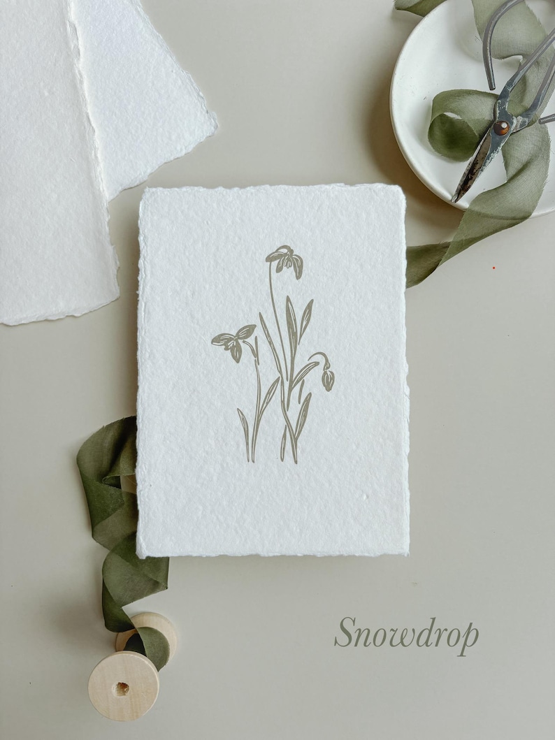 May include: A white, textured card with a botanical illustration of snowdrop flowers in a muted green hue. The word "Snowdrop" is printed in a matching color. A green ribbon, scissors, and additional paper are arranged around the card.