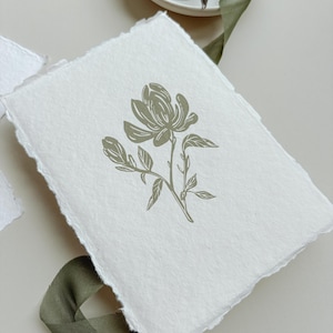 May include: A white handmade paper card with a torn edge, featuring a sage green floral illustration of a magnolia flower. The card is placed on a neutral surface with a green ribbon and a small white dish with scissors.