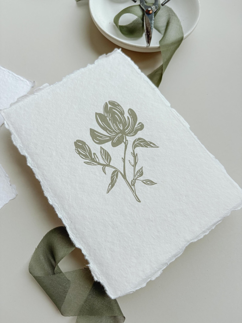 May include: A white handmade paper card with a torn edge, featuring a sage green floral illustration of a magnolia flower. The card is placed on a neutral surface with a green ribbon and a small white dish with scissors.