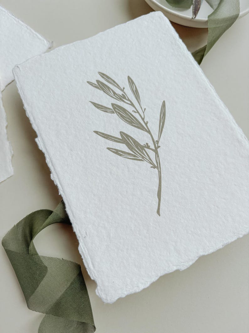 May include: A white, textured paper card with a torn edge, featuring a muted green illustration of an olive branch. A green ribbon is draped nearby. The card is likely for stationery or invitations.