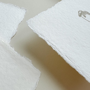 May include: Close-up of two pieces of handmade, off-white paper with deckled edges. One piece has a delicate floral design in a muted gray tone. The paper has a textured surface and a soft, natural appearance, suggesting a focus on stationery or art supplies.