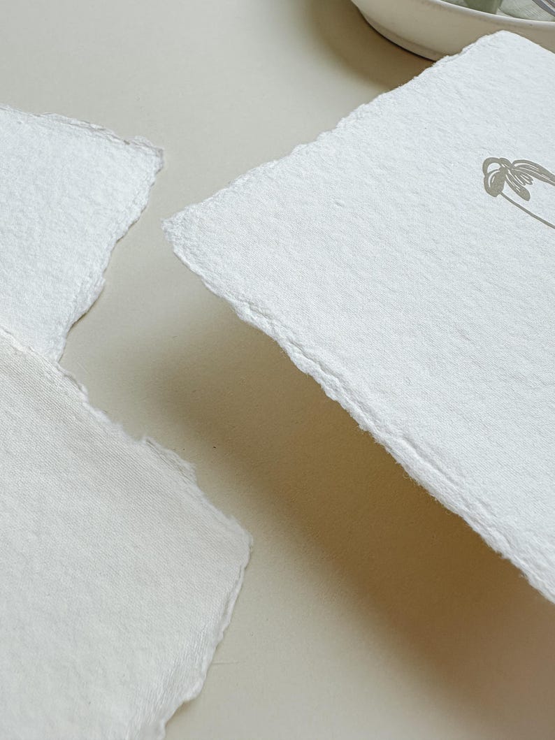 May include: Close-up of two pieces of handmade, off-white paper with deckled edges. One piece has a delicate floral design in a muted gray tone. The paper has a textured surface and a soft, natural appearance, suggesting a focus on stationery or art supplies.