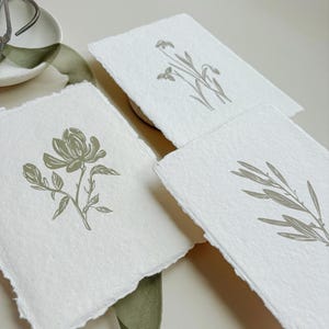 May include: Three handmade paper cards with botanical illustrations. Each card features a different design: a flower, a sprig, and a branch. The illustrations are in a muted green color, and the paper has a deckled edge.