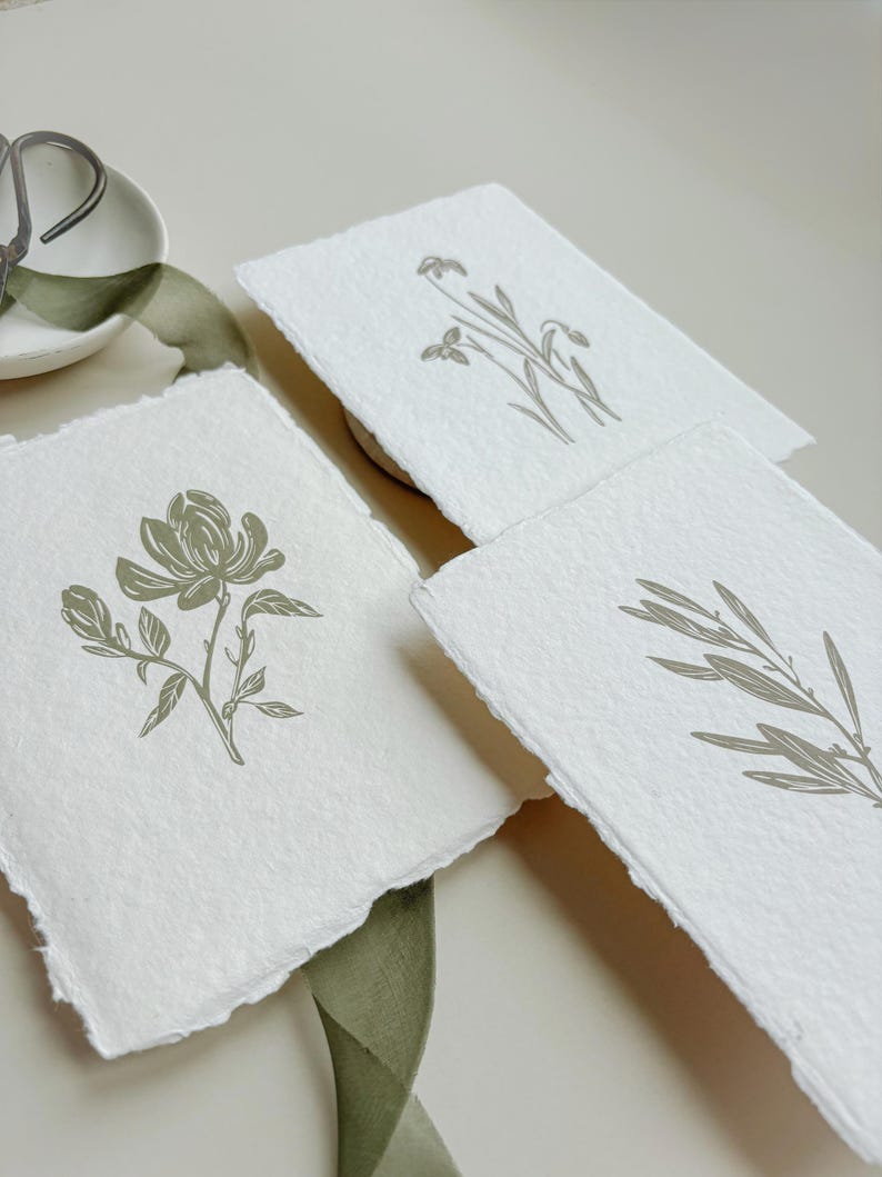 May include: Three handmade paper cards with botanical illustrations. Each card features a different design: a flower, a sprig, and a branch. The illustrations are in a muted green color, and the paper has a deckled edge.