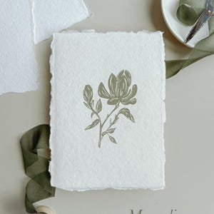 May include: A handmade paper card with a magnolia flower illustration in olive green. The card has a deckled edge and the word "Magnolia" is printed at the bottom. A green ribbon and a wooden spool are visible.