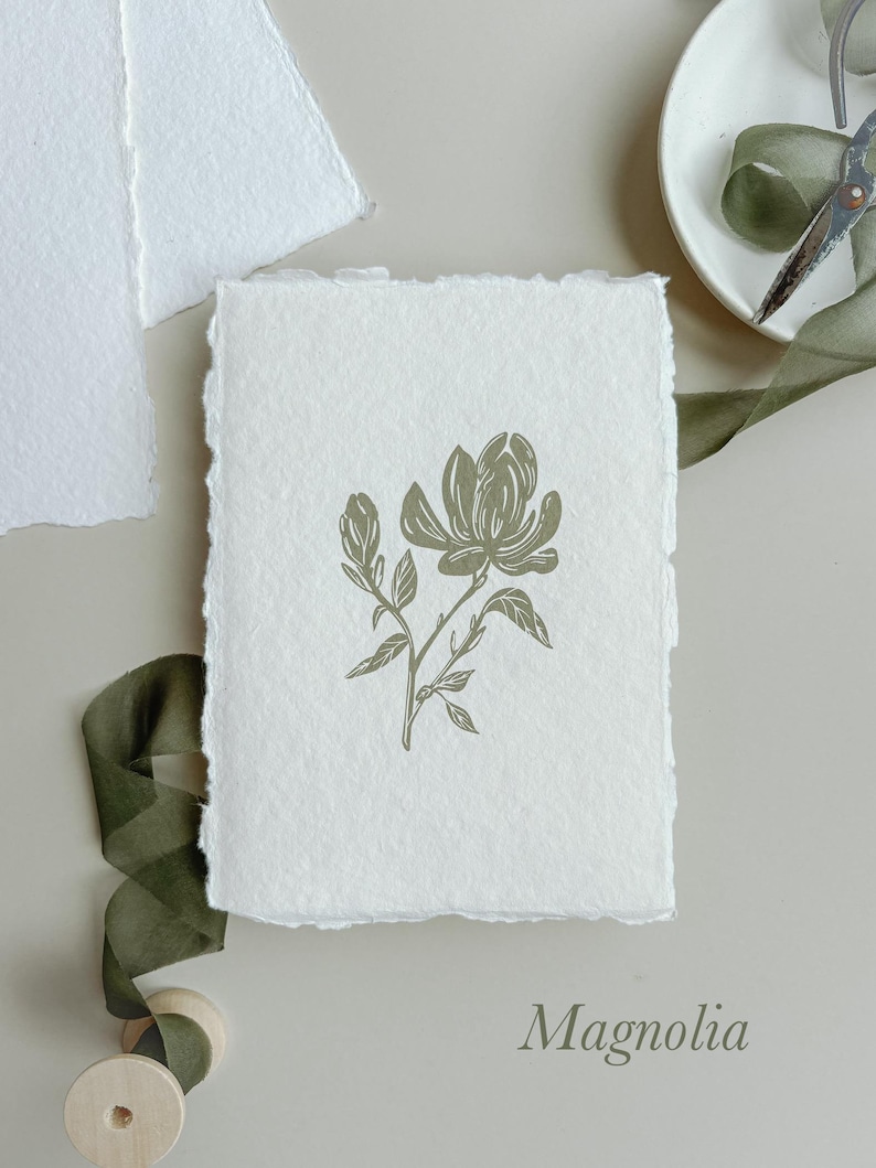 May include: A handmade paper card with a magnolia flower illustration in olive green. The card has a deckled edge and the word "Magnolia" is printed at the bottom. A green ribbon and a wooden spool are visible.