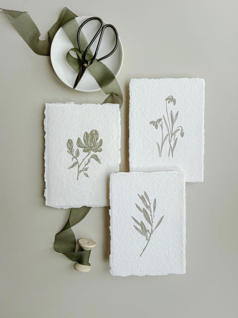 May include: Three botanical art prints with olive green illustrations of flowers and leaves on textured white paper. A white plate with black scissors and olive green ribbon are also in the image. A spool of olive green thread is also visible.