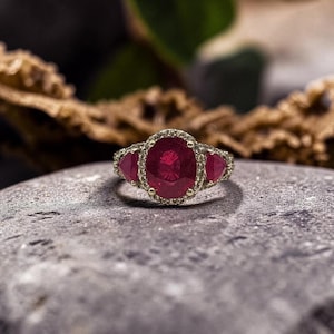 May include: A gold ring featuring a large oval-cut red ruby center stone surrounded by a halo of small white diamonds. Two triangular-cut red rubies are set on either side of the center stone, all mounted on a thin gold band with a polished finish.