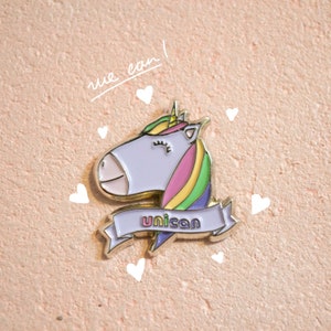 Może przedstawiać: Złota szpilka emaliowana z kreskówkowym jednorożcem z tęczową grzywą i banerem z napisem "unicon".