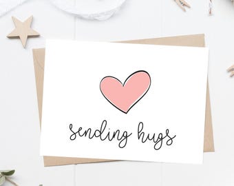 May include: A white greeting card featuring a hand-drawn style pink heart with a black outline centered above the black cursive text that reads sending hugs. The card is placed on top of a brown envelope, creating a simple and clean visual presentation.