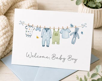 Op de afbeelding: Een witte wenskaart met de tekst Welcome, Baby Boy in zwarte schrijfletters. De illustratie toont een waslijn met een rompertje met sterren, blauwe sokjes, een blauwe slaapzak, een groene tuinbroek, een blauw t-shirt en een blauwe teddybeer.