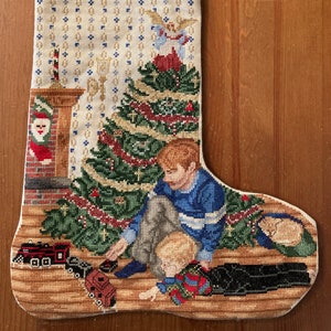 May include: A white Christmas stocking with a red and black train at the top, the name "DANIEL" embroidered in green, and a scene of two children playing with toys in front of a Christmas tree.