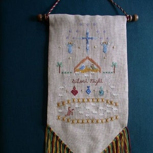 May include: A beige linen tapestry with a cross, stars, and a nativity scene. The tapestry has a fringe border and the text "Silent Night" is embroidered in the centre.