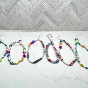 Può includere: Otto ciondoli per telefono colorati con perline, con diversi design e lettere. I ciondoli sono realizzati con piccole perline colorate e includono lettere, faccine sorridenti, stelle e altre forme.