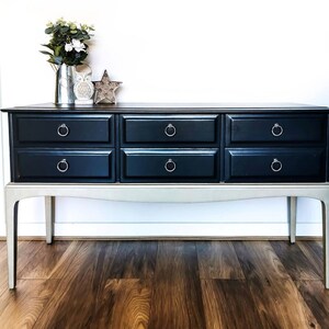 May include: A black dresser with six drawers and silver ring pulls. The dresser has a light grey base and sits on four legs. The dresser is in front of a white wall and a wooden floor.