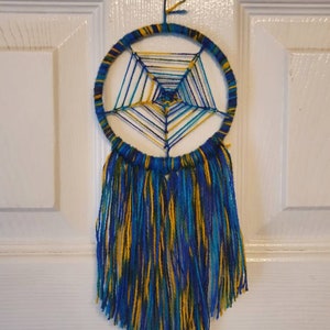 Puede incluir: Un atrapasue&ntilde;os hecho a mano con un marco circular envuelto en hilo azul y amarillo. La red interior est&aacute; tejida con hilos azules, amarillos y turquesas. Largas y fluidas borlas en colores a juego cuelgan debajo, creando una decoraci&oacute;n de pared.