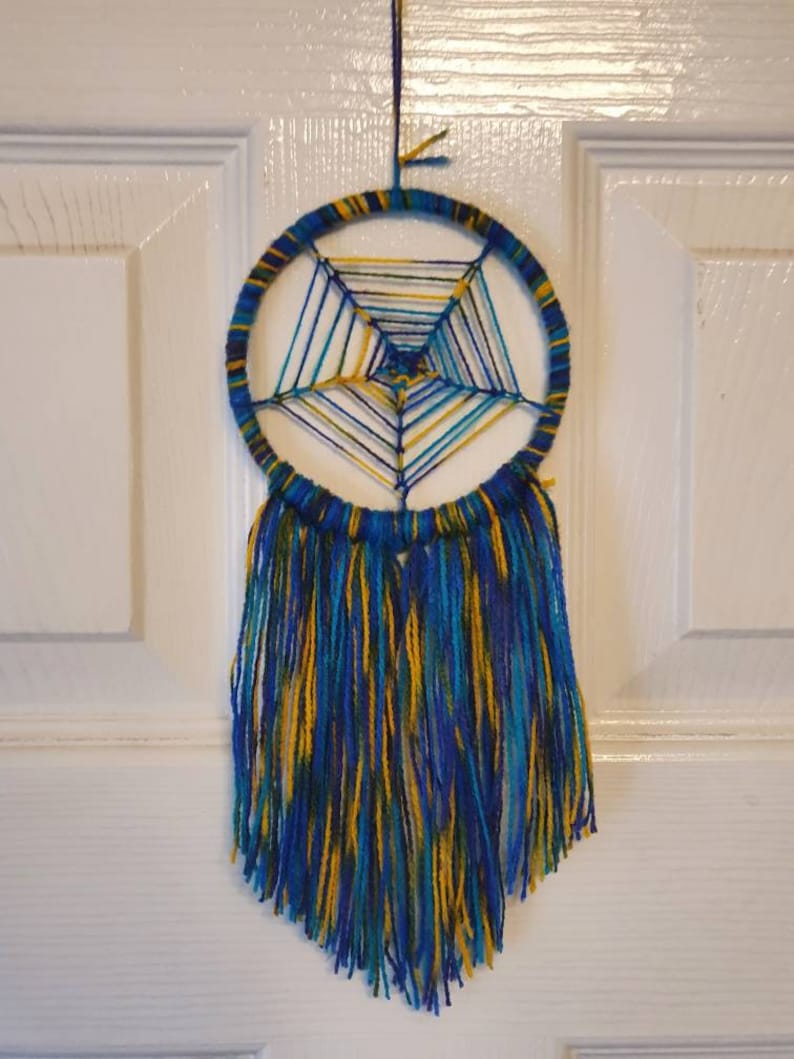 Puede incluir: Un atrapasue&ntilde;os hecho a mano con un marco circular envuelto en hilo azul y amarillo. La red interior est&aacute; tejida con hilos azules, amarillos y turquesas. Largas y fluidas borlas en colores a juego cuelgan debajo, creando una decoraci&oacute;n de pared.
