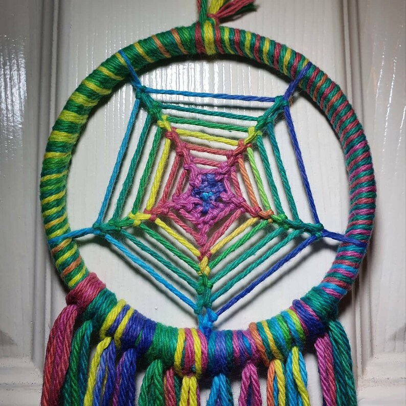 Puede incluir: Un atrapasue&ntilde;os hecho a mano con un marco circular envuelto en hilo verde, amarillo y naranja. La red est&aacute; tejida con hilos de colores del arco&iacute;ris y borlas coloridas cuelgan de la parte inferior. Dise&ntilde;ado para colgar.