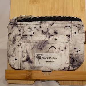Pode incluir: Uma bolsa retangular de tecido com um padrão de fumaça em preto e branco, estrelas celestiais e luas crescentes. Possui um fecho de correr preto, uma aba lateral castanha com um anel de metal e uma etiqueta branca com o texto impresso.