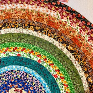 May include: A circular fabric mat constructed from concentric rings of various floral-patterned cotton strips in a rainbow color gradient, ranging from cream and blue to green, orange, and red, featuring a small pink tag labeled Handmade.