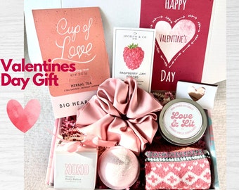 May include: A pink and white Valentine's Day gift box filled with various items, including a pink hair scrunchie, a bath bomb, a candle, a box of tea, a jar of raspberry jam, and a pair of pink and white socks. The box also includes a card that says "Happy Valentine's Day".