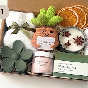 May include: A gift box filled with self-care items. Includes a white towel with a green ribbon, a crochet succulent, a candle with star anise, a lip balm, a face mask, a green soap bar, and a flower hair clip.