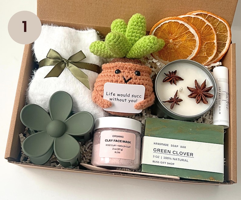 May include: A gift box filled with self-care items. Includes a white towel with a green ribbon, a crochet succulent, a candle with star anise, a lip balm, a face mask, a green soap bar, and a flower hair clip.