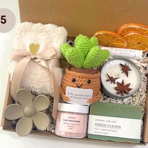 May include: A gift box filled with self-care items. Includes a cream-colored towel with a gold heart, a crochet cactus, a candle, soap, clay face mask, and dried orange slices. The box contains the text "Life would succ without you!"