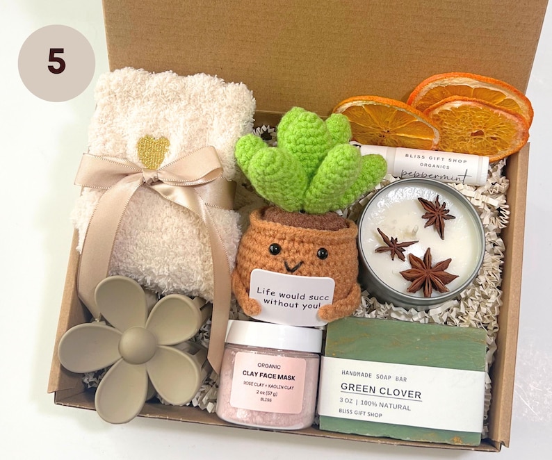 May include: A gift box filled with self-care items. Includes a cream-colored towel with a gold heart, a crochet cactus, a candle, soap, clay face mask, and dried orange slices. The box contains the text "Life would succ without you!"