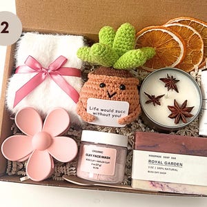 May include: A gift box filled with self-care items. Includes a white fluffy towel with a pink ribbon, a crochet cactus, a candle with star anise, a lip balm, a pink flower, a face mask, and a bar of soap.