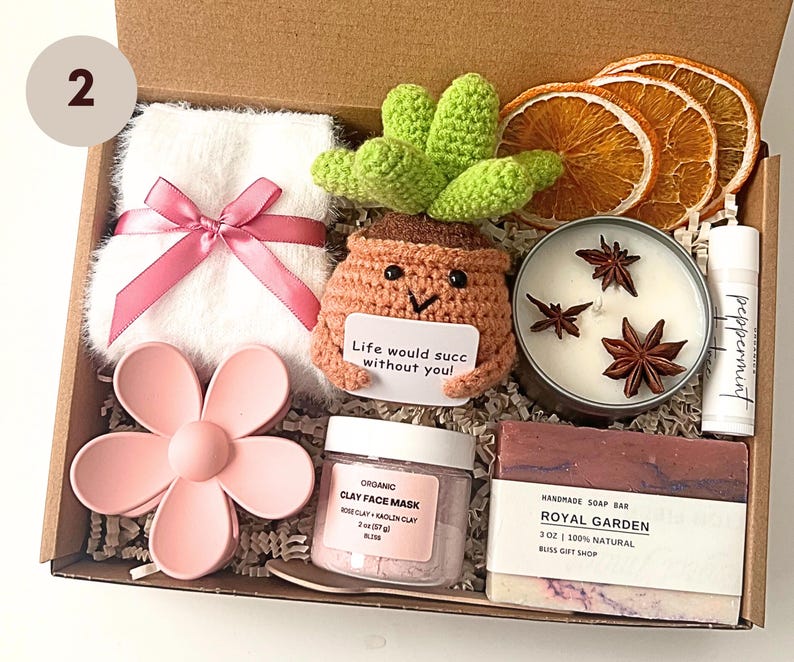 May include: A gift box filled with self-care items. Includes a white fluffy towel with a pink ribbon, a crochet cactus, a candle with star anise, a lip balm, a pink flower, a face mask, and a bar of soap.