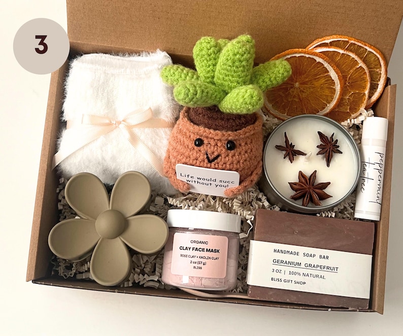 May include: A gift box filled with self-care items. Includes a white fluffy item with a bow, a crocheted cactus in a pot, a candle with star anise, a lip balm, a flower-shaped item, a jar of clay face mask, and a bar of soap.