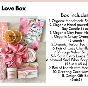 May include: A pink and white gift box filled with various items, including a pink scrunchie, a clay face mask, a soy candle, a card that says "Happy Mother's Day", and a soap saver bag. The box includes a variety of other items, including herbal tea, orange slices, and chenille socks.
