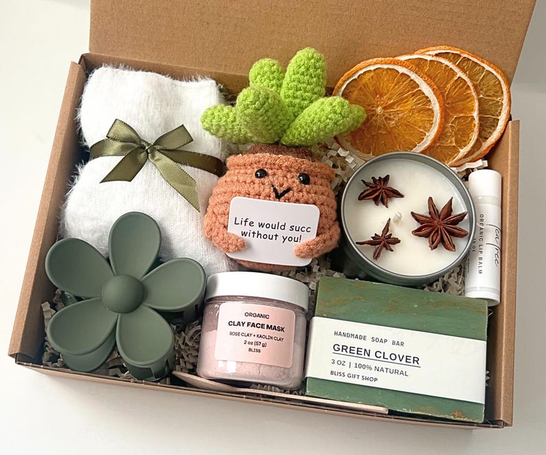 May include: A gift box filled with self-care items. Includes a white towel with a green ribbon, a green flower hair clip, a crochet succulent, a candle, lip balm, clay face mask, soap bar, and dried orange slices.