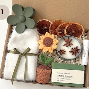 K&ouml;nnte beinhalten: Eine Geschenkbox gef&uuml;llt mit verschiedenen Artikeln, darunter eine geh&auml;kelte Sonnenblume, eine gr&uuml;ne Seife, eine Kerze mit Sternanis, ein wei&szlig;er flauschiger Schal, eine gr&uuml;ne Blumenhaarklammer und ein Teebaum-Lippenbalsam. Die Seife ist mit "Handmade Soap Bar Green Clover 3 oz 100% Natural Bliss Gift Shop" beschriftet.