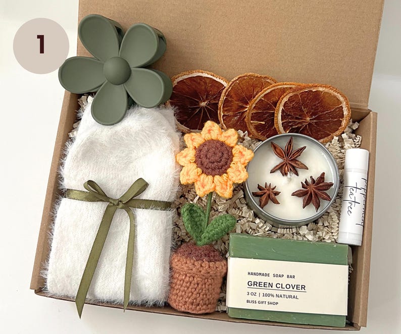 K&ouml;nnte beinhalten: Eine Geschenkbox gef&uuml;llt mit verschiedenen Artikeln, darunter eine geh&auml;kelte Sonnenblume, eine gr&uuml;ne Seife, eine Kerze mit Sternanis, ein wei&szlig;er flauschiger Schal, eine gr&uuml;ne Blumenhaarklammer und ein Teebaum-Lippenbalsam. Die Seife ist mit "Handmade Soap Bar Green Clover 3 oz 100% Natural Bliss Gift Shop" beschriftet.
