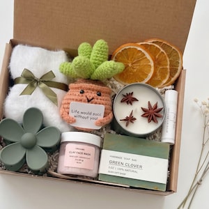 May include: A gift box filled with self-care items. Includes a white towel with a green ribbon, a crochet succulent, a candle, a face mask, a soap bar, and dried orange slices. The box also contains a green flower hair clip and lip balm.