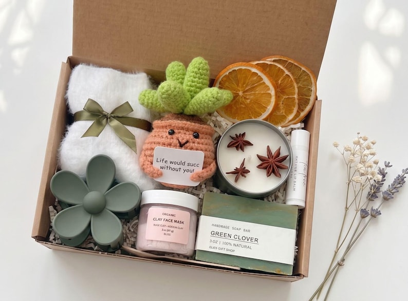 May include: A gift box filled with self-care items. Includes a white towel with a green ribbon, a crochet succulent, a candle, a face mask, a soap bar, and dried orange slices. The box also contains a green flower hair clip and lip balm.