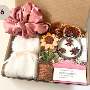 K&ouml;nnte beinhalten: Eine Geschenkbox gef&uuml;llt mit verschiedenen Artikeln, darunter ein rosa Satin-Haargummi, eine wei&szlig;e, flauschige Augenmaske, eine geh&auml;kelte Sonnenblume, eine kleine Topfpflanze, eine Kerze mit Sternanis, eine Seife mit der Aufschrift "Handmade Soap Bar Geranium Grapefruit 3 oz | 100% Natural Bliss Gift Shop" und eine kleine Flasche mit der Aufschrift "tea tree".
