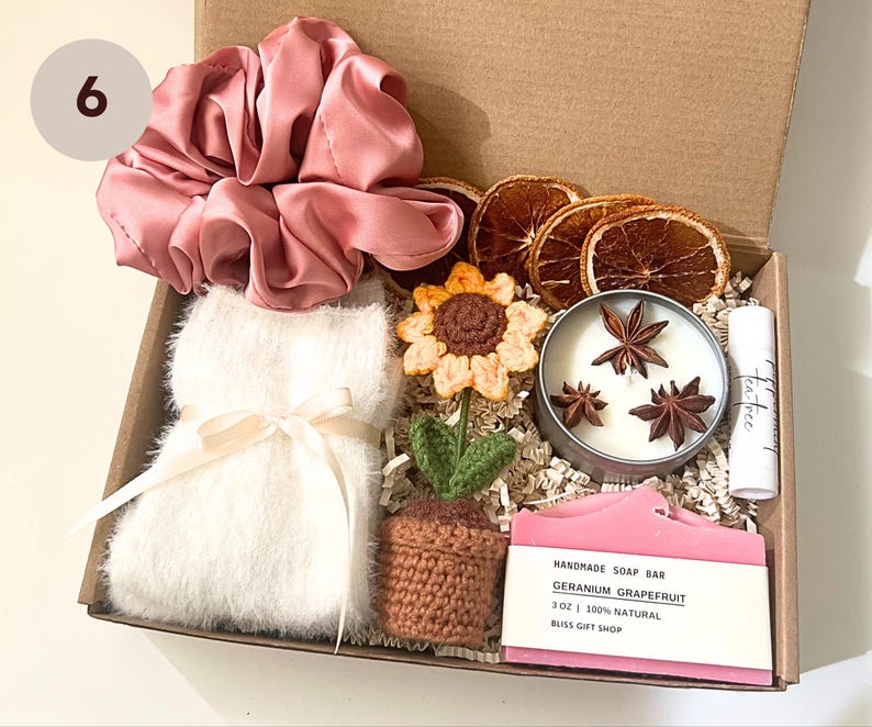 K&ouml;nnte beinhalten: Eine Geschenkbox gef&uuml;llt mit verschiedenen Artikeln, darunter ein rosa Satin-Haargummi, eine wei&szlig;e, flauschige Augenmaske, eine geh&auml;kelte Sonnenblume, eine kleine Topfpflanze, eine Kerze mit Sternanis, eine Seife mit der Aufschrift "Handmade Soap Bar Geranium Grapefruit 3 oz | 100% Natural Bliss Gift Shop" und eine kleine Flasche mit der Aufschrift "tea tree".