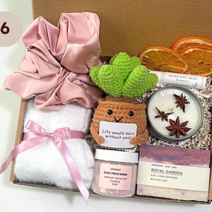 May include: A gift box filled with self-care items. Includes a pink satin scrunchie, a white towel with a pink ribbon, a crocheted succulent, a candle, a bar of soap, and a jar of clay face mask. The box contains the text "Life would succ without you!"