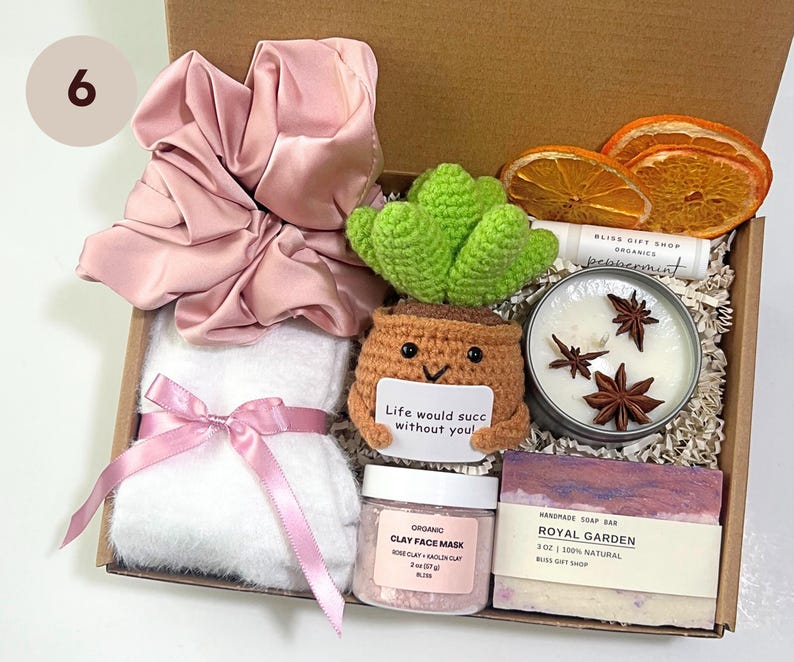 May include: A gift box filled with self-care items. Includes a pink satin scrunchie, a white towel with a pink ribbon, a crocheted succulent, a candle, a bar of soap, and a jar of clay face mask. The box contains the text "Life would succ without you!"