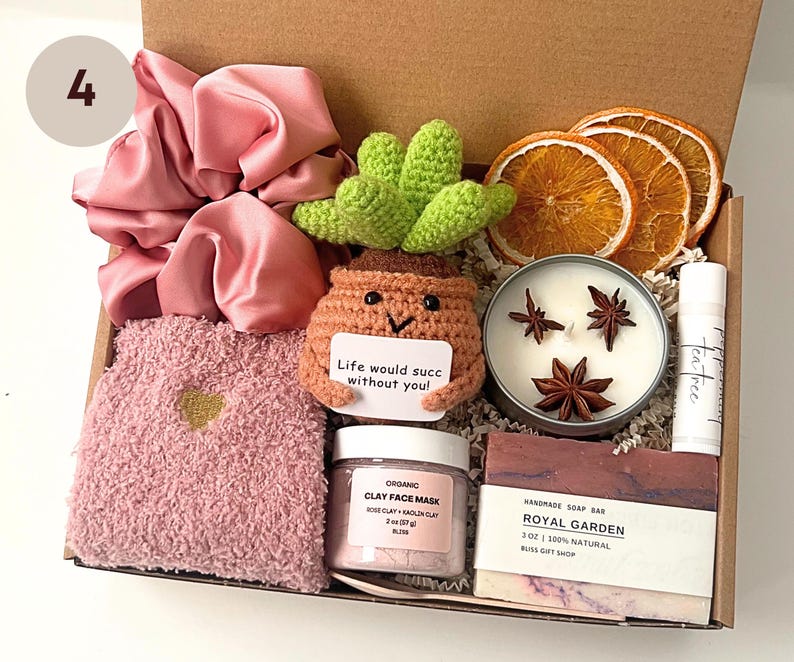 May include: A gift box filled with self-care items. Includes a pink scrunchie, fuzzy socks, a crocheted succulent, a candle, lip balm, a face mask, and a bar of soap. The succulent has a tag that reads "Life would succ without you!"