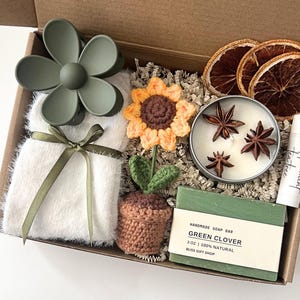 K&ouml;nnte beinhalten: Eine Geschenkbox gef&uuml;llt mit verschiedenen Artikeln, darunter eine geh&auml;kelte Sonnenblume, eine gr&uuml;ne Kleeblattseife, eine Kerze mit Sternanis, ein wei&szlig;er flauschiger Schal, eine gr&uuml;ne Blumenhaarklammer und eine kleine geh&auml;kelte Topfpflanze. Die Box ist mit braunem zerkleinertem Papier gef&uuml;llt.