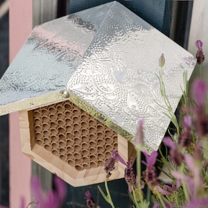 May include: A hexagonal wooden bee house with a silver embossed metal roof. The bee house is mounted on a wall and surrounded by lavender plants.