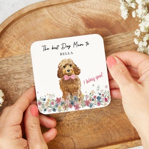 May include: Square coaster featuring a watercolour illustration of a brown dog with a pink bow tie, surrounded by flowers. Text reads "The best Dog Mum to BELLA" and "I Woof you!"